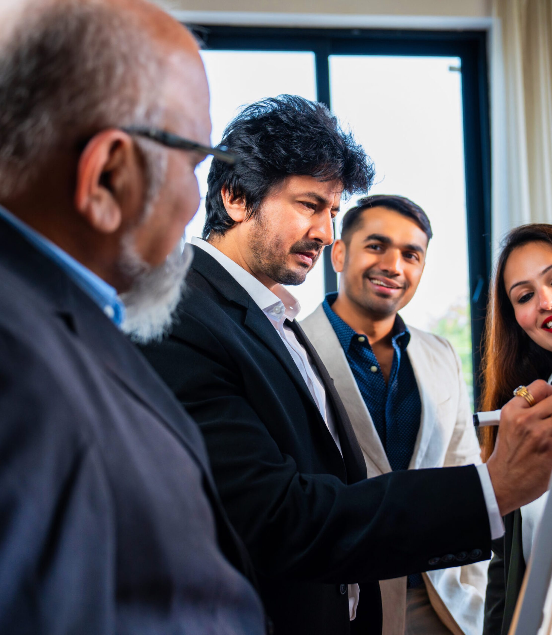 Indian Asian young business professional or businessman explaining strategy on a whiteboard while colleagues stand nearby observing and discussing ideas in a modern corporate office, side angle view
